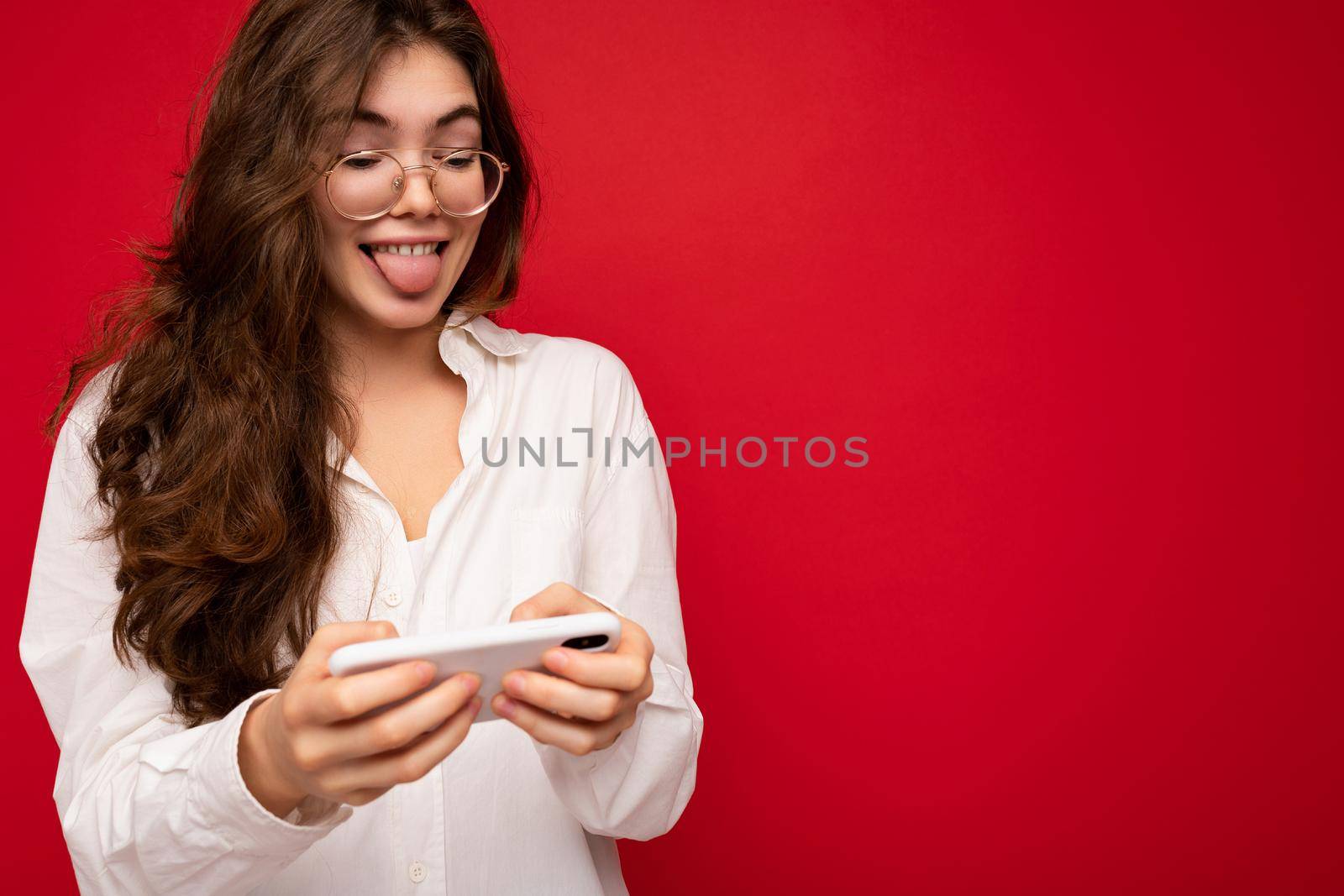 Portrait photo of Pretty smiling positive young brunette woman wearing white shirt and optical glasses isolated over red background holding in hand and using mobile phone playing online games looking at gadjet display and showing tongue.