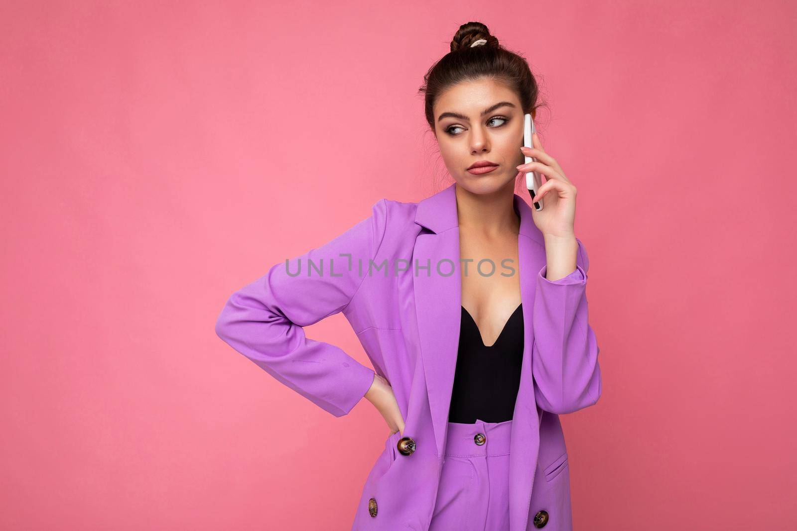 Photo of dissatisfied beautiful young woman wearing purple suit talking on the mobile phone isolated over pink background looking to the side.