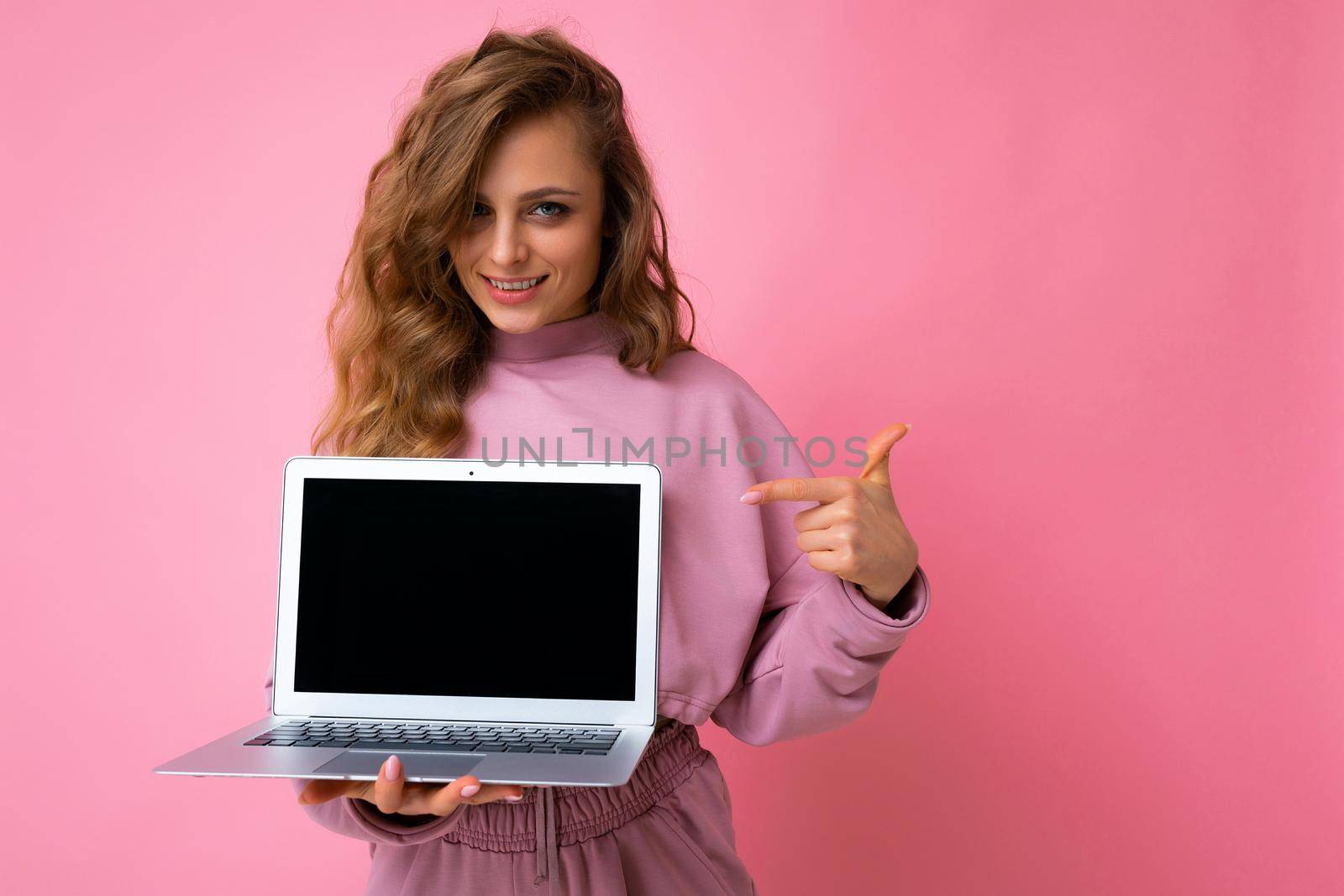 Photo of beautiful blond curly young woman lookind at camera holding computer laptop with empty monitor screen with mock up and copy space pointing at netbook with hand and one finger wearing pink hoodie isolated over pink wall background.
