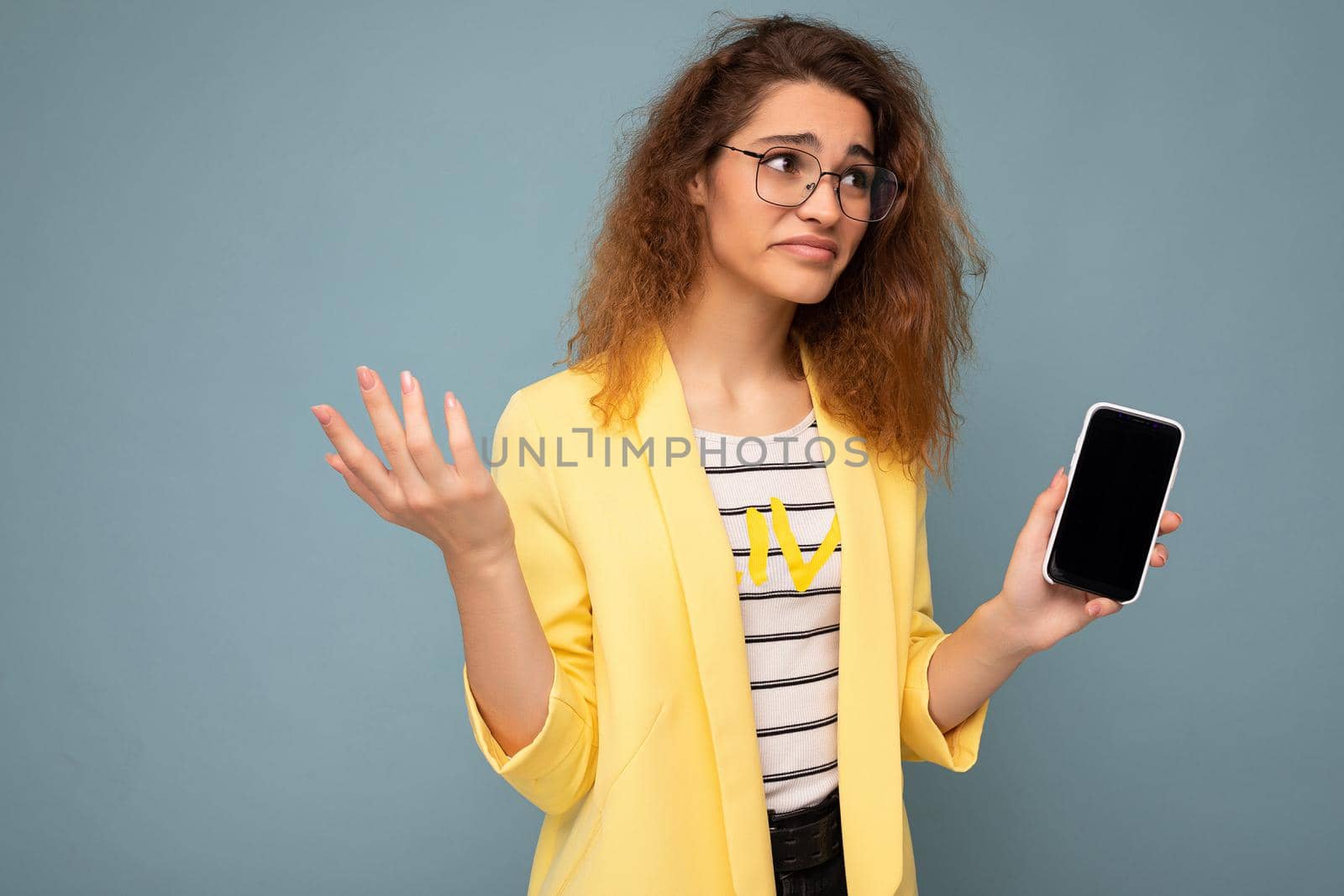 attractive asking young woman with curly dark blond hair wearing yellow jacket and optical glasses isolated on background holding and showing mobile phone with empty space for cutout looking to the side and having doubts.