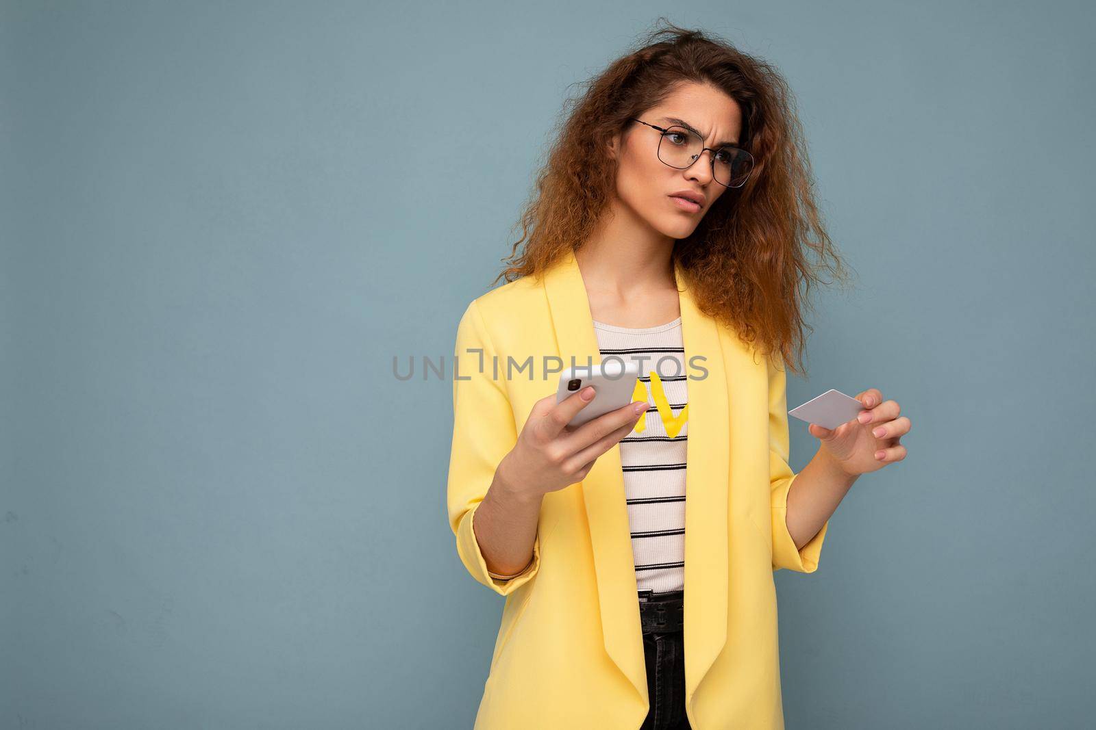 Pretty upset sad woman wearing everyday stylish clothes isolated on background wall holding and using phone and credit card making payment looking to the side.