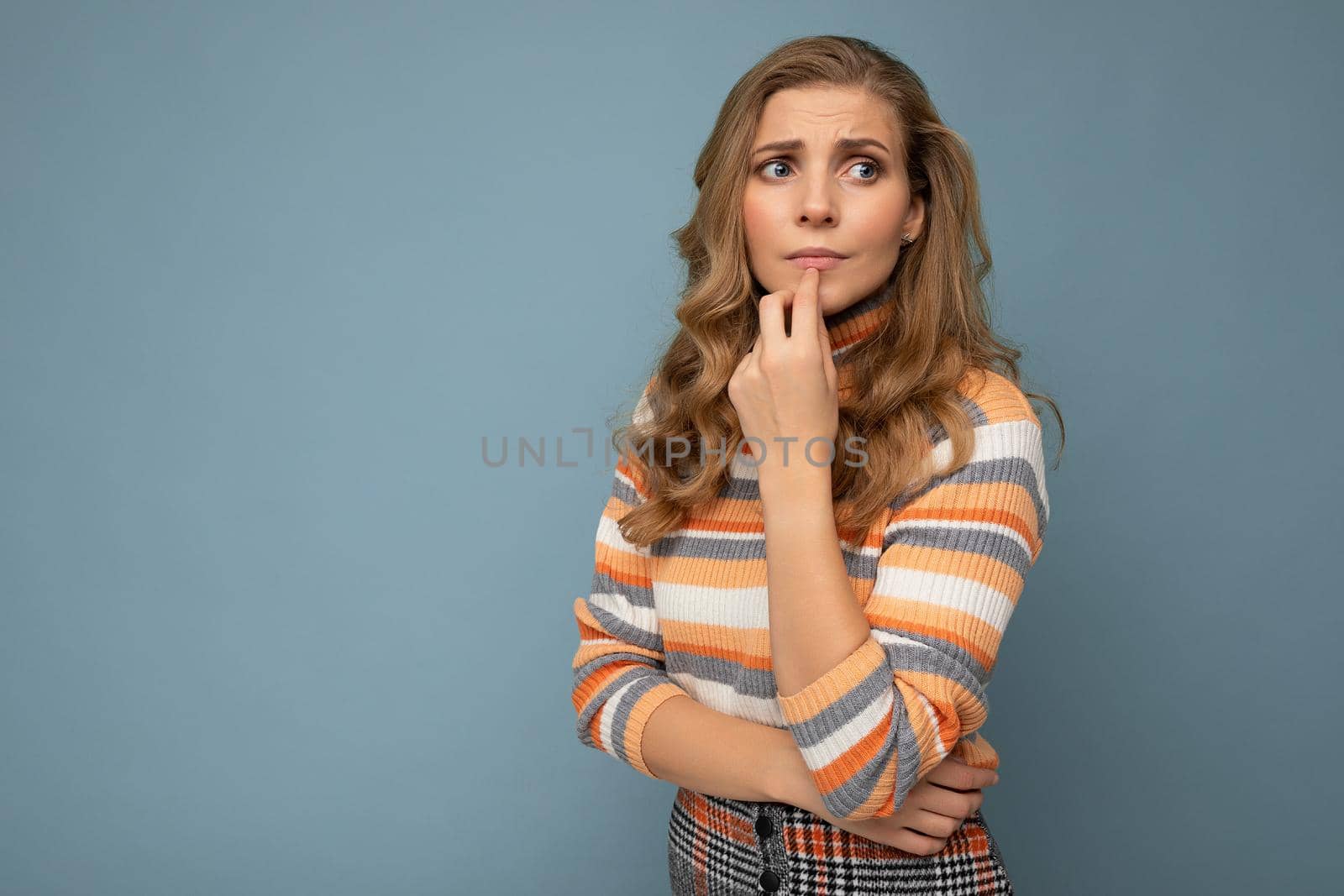 Photo portrait of young attractive beautiful sad upset thoughtful blonde woman with sincere emotions wearing striped sweater isolated on blue background with empty space and has doubts.