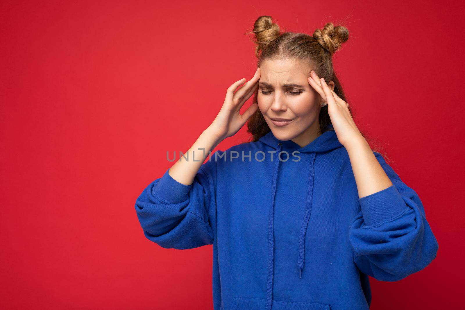 Photo of tired weary young nice pretty blond woman with two horns with sincere emotions wearing casual bright blue hoodie isolated over red background with copy space and having headache. Pain concept.