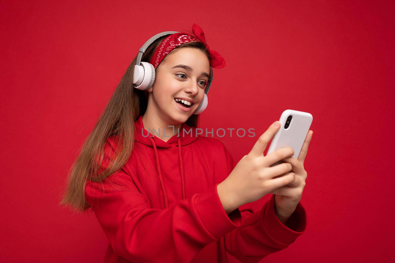 Positive attractive brunette girl wearing red hoodie isolated on red background holding and using smartphone cwatching videos online wearing white wireless headphones listening to cool music looking at gadjet display.