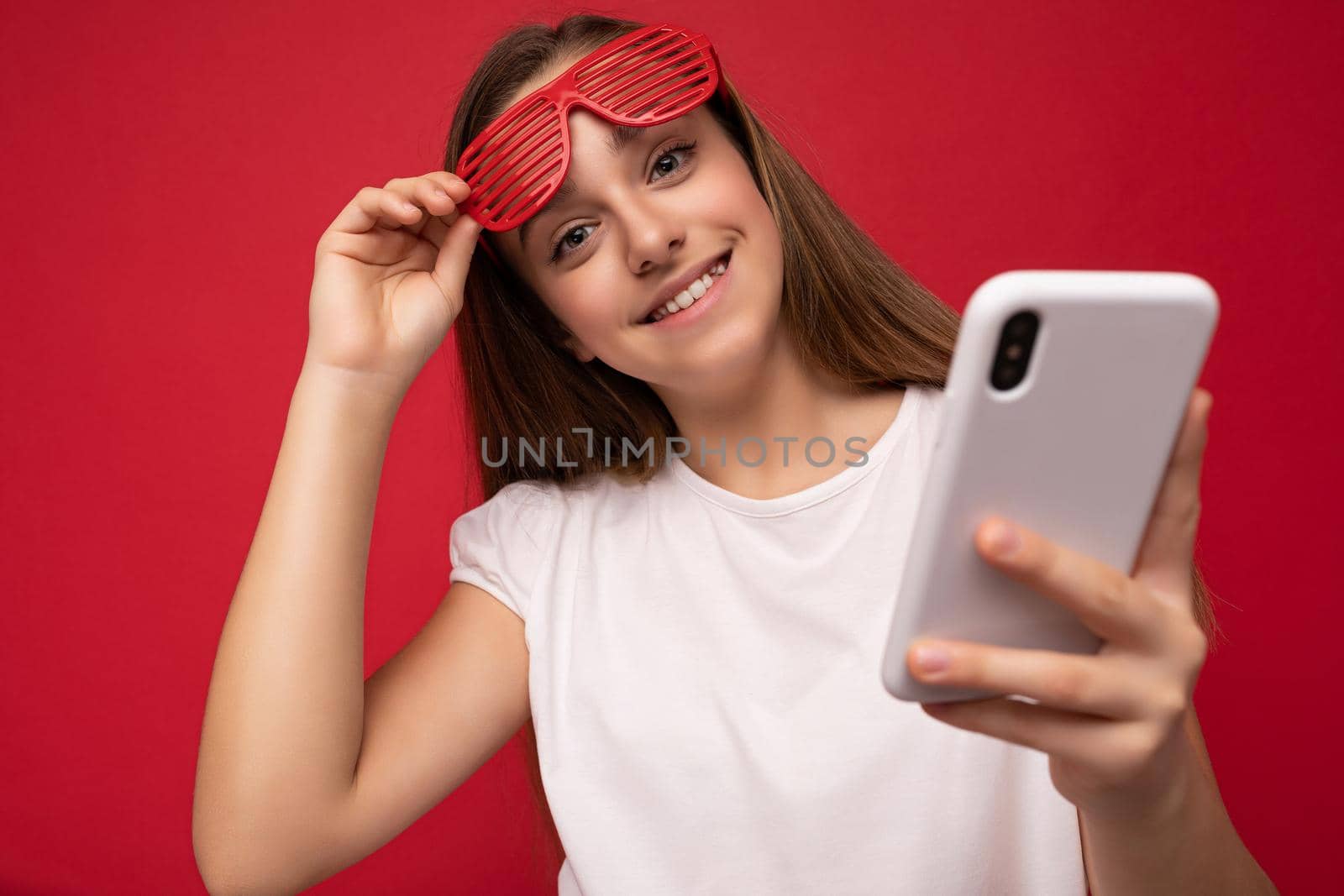 Charming positive young girl wearing white t-shirt and red glasses poising isolated on red background with empty space holding in hand and using mobile phone communicating online looking at camera.