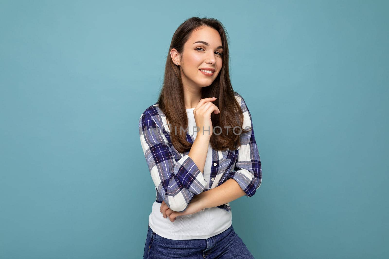 Young positive smiling beautiful brunette woman with sincere emotions wearing trendy check shirt standing isolated on blue background with empty space.