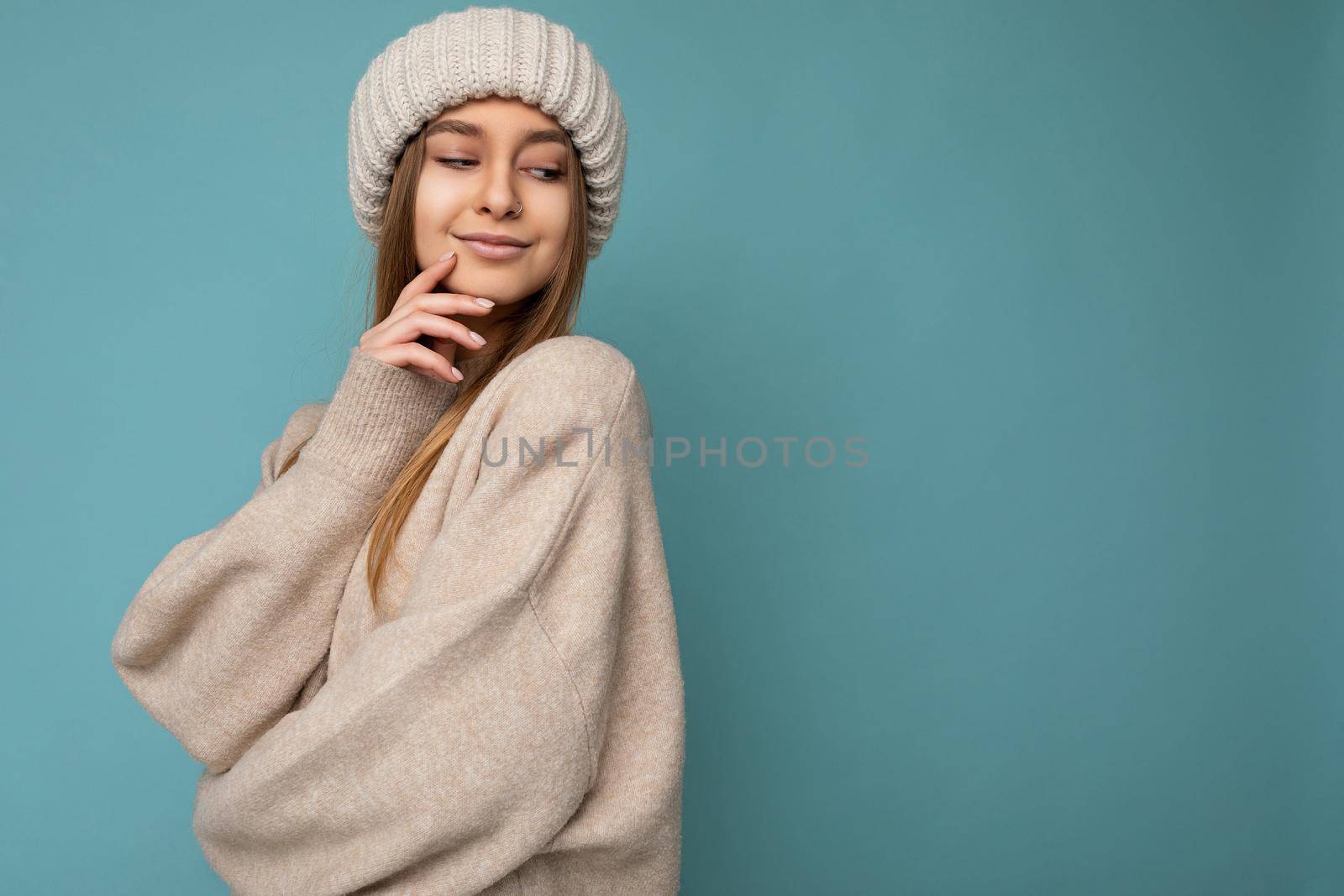 Shot of sexy pretty happy young dark blonde woman standing isolated on blue background wall wearing beige warm sweater and winter beige hat looking down. Free space, copy space