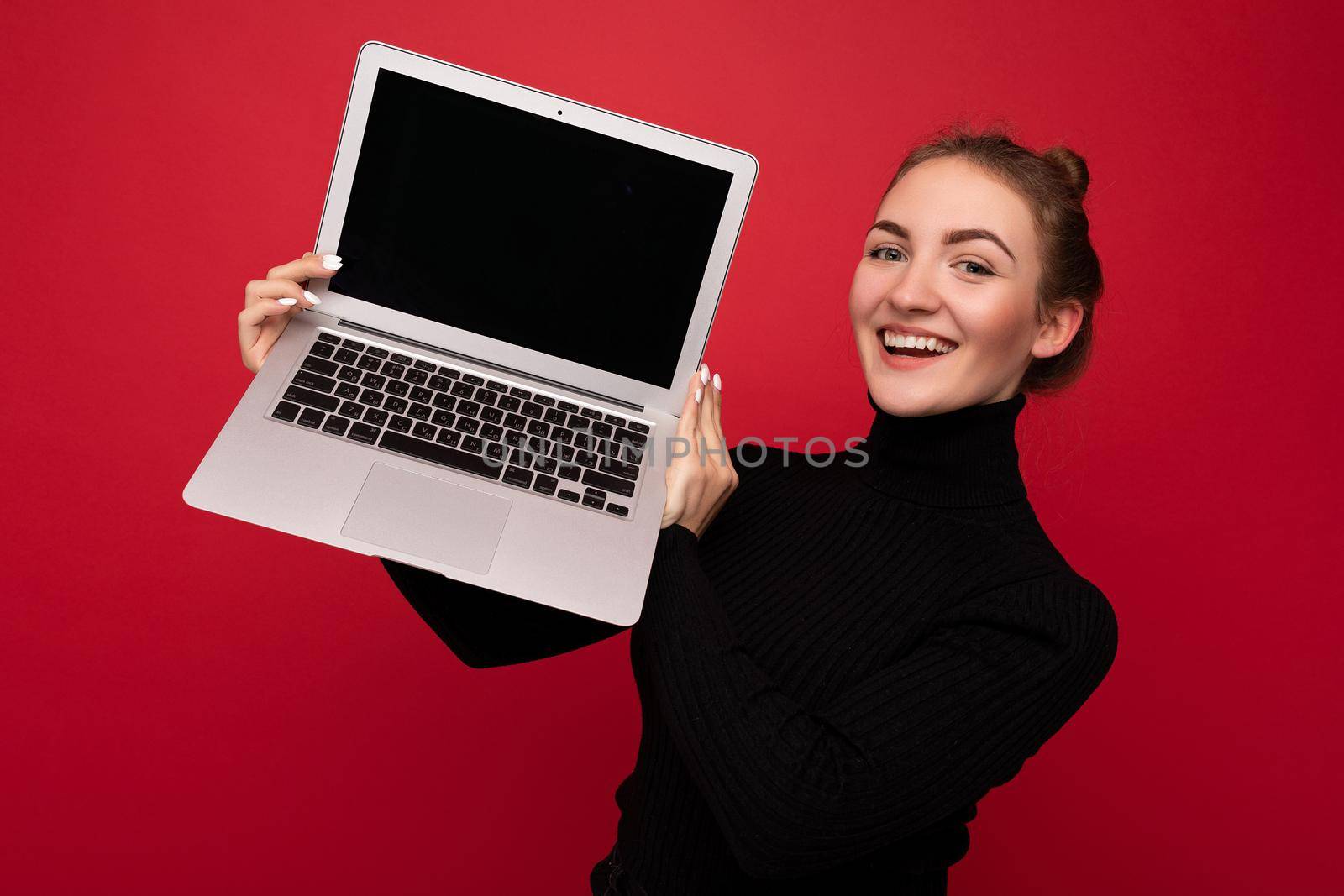 Close-up portrait of Beautiful smiling happy young woman with gathered hair holding computer laptop with empty monitor screen with mock up wearing black longsleeve looking at camera.