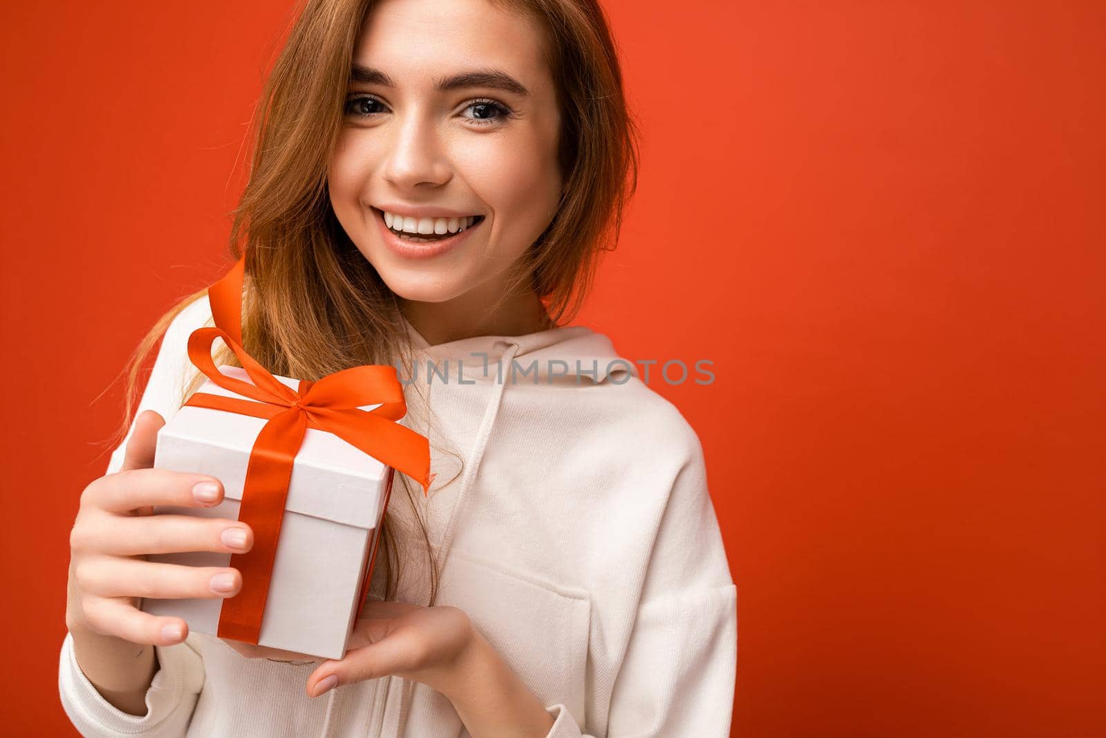 Beautiful happy young blonde woman isolated over colourful background wall wearing stylish casual clothes holding gift box and looking at camera. Free space