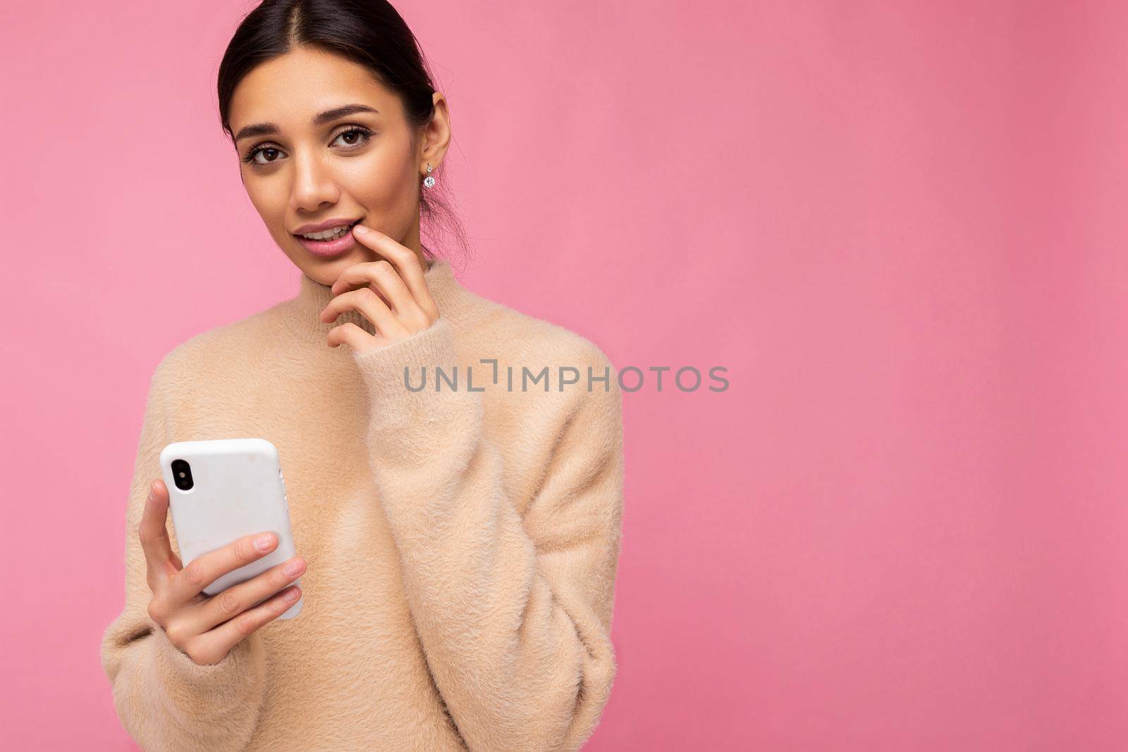 Closeup photo of Attractive cute young brunet woman wearing beige warm sweater standing isolated over pink background surfing on the internet via phone looking at camera and being concerned.