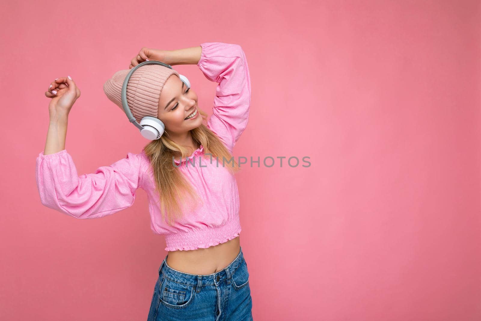 Beautiful happy smiling young blonde woman wearing pink blouse and pink hat isolated over pink background wall wearing white wireless bluetooth headphones listening to cool music having fun and dancing. copy space