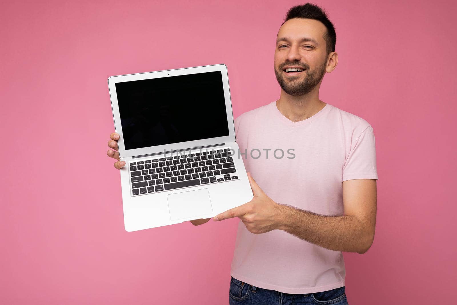 Handsome smiling brunet man holding laptop computer looking at camera in t-shirt on isolated pink background.