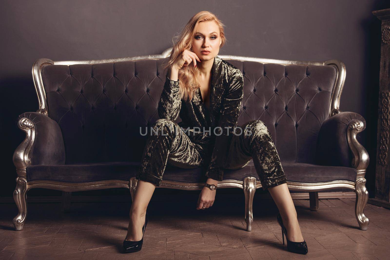 Beautiful young woman in a velor suit sits on a gray vintage couch.