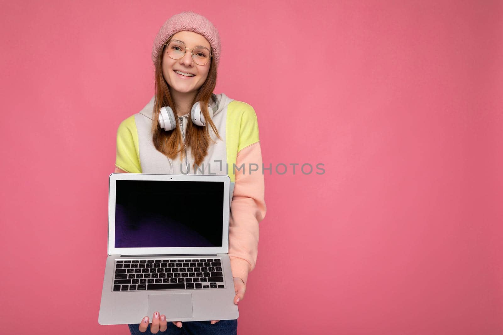 Pretty Smiling happy female teenager wearing hat colorful sweater and glasses holding computer laptop with empty copy space screen monitor wearing white headphones looking at camera isolated on pink wall background. Mock up