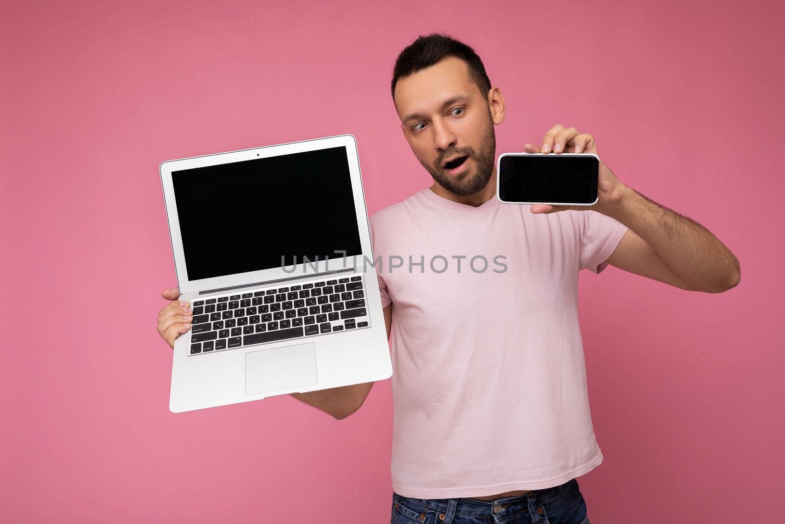 Handsome brunet man with open mouth holding laptop computer and mobile phone looking at phone in t-shirt on isolated pink background.