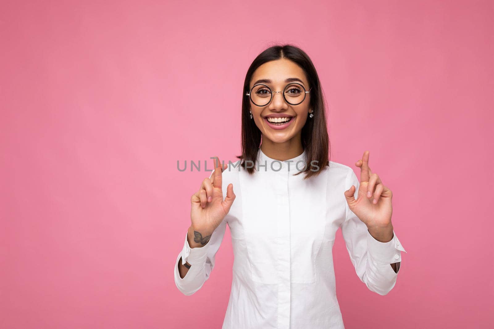 Portrait of young positive happy attractive brunette woman with sincere emotions wearing casual white shirt and optical glasses isolated over pink background with copy space and holding fingers crossed for good luck.