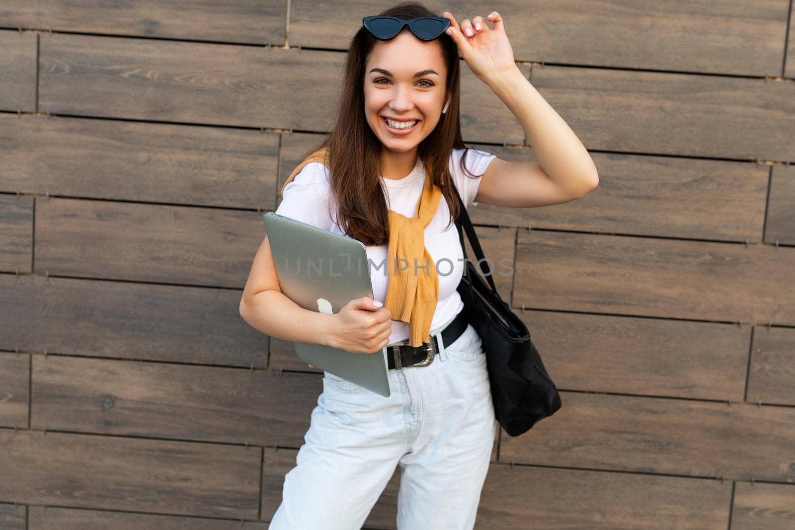 Photo of beautiful fascinating charming smiling young brunette girl wearing white t-shirt light blue jeans and yellow sweater holding computer laptop and black sunglasses staying at the street and looking at camera.