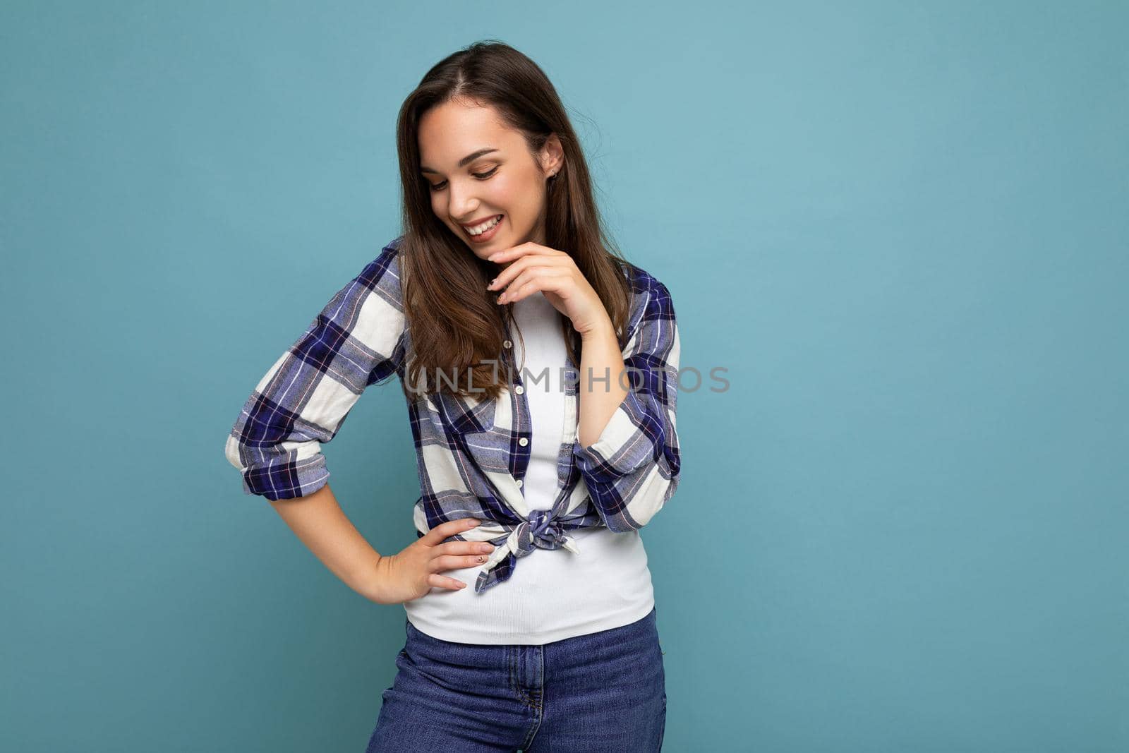 Photo portrait of young beautiful smiling hipster brunette woman in trendy blue and white shirt and jeans clothes. Sexy carefree female person posing isolated near blue wall with empty space in studio. Positive model with natural makeup by TRMK
