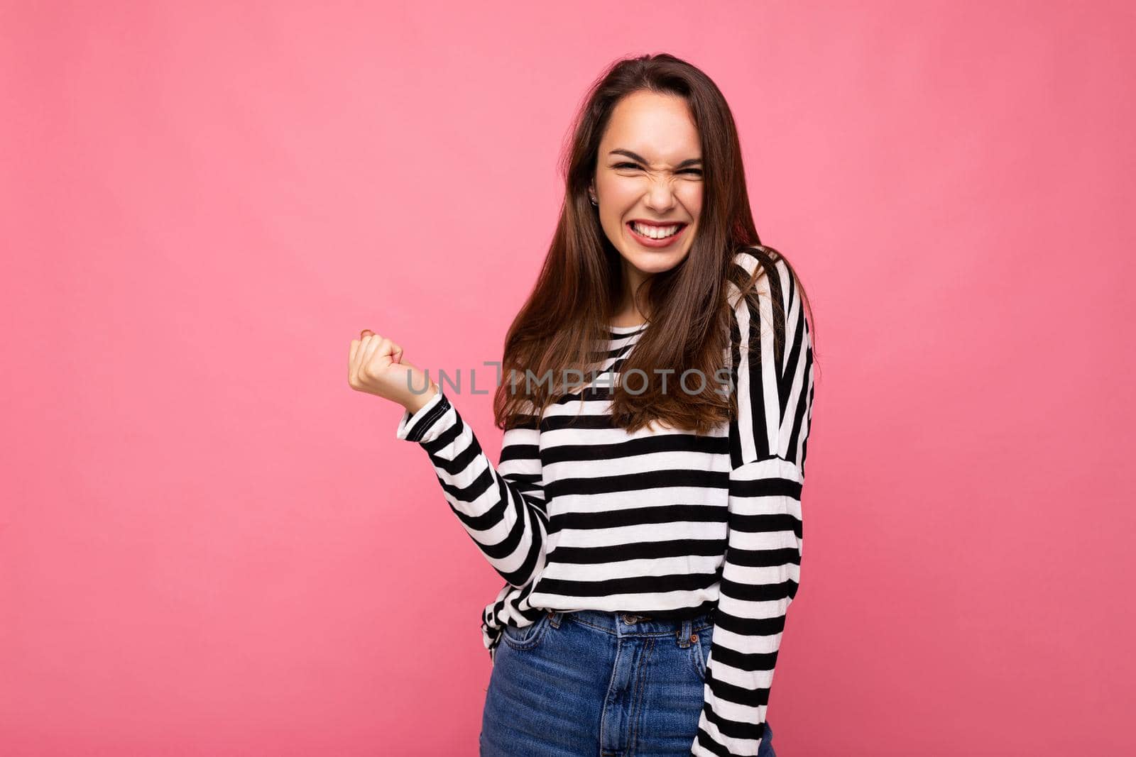 Portrait of young positive happy attractive brunette woman with sincere emotions in casual striped pullover isolated on pink background with free space and celebrating winning shouting yeah.