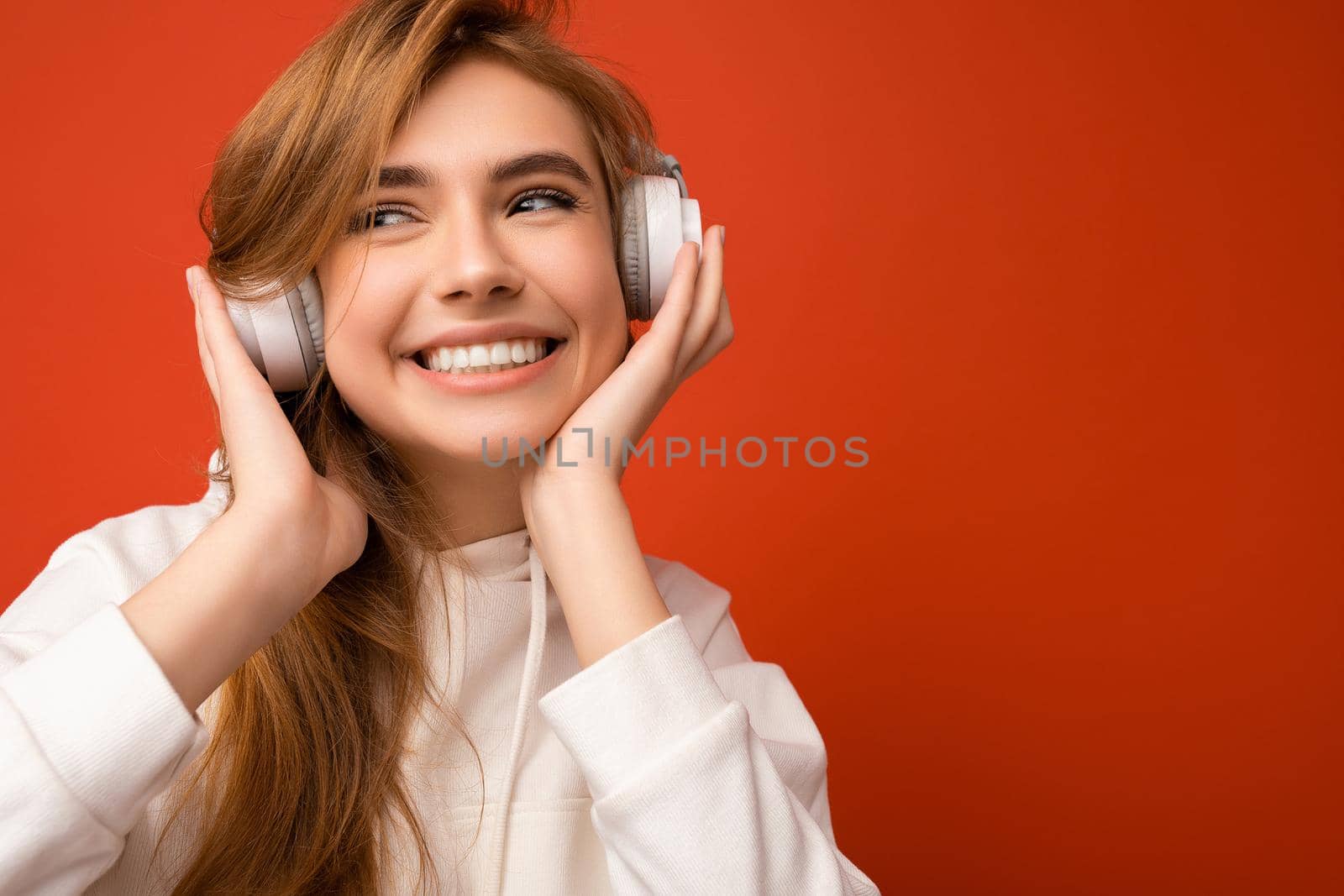 Closeup photo of attractive positive young blonde woman wearing white hoodie isolated over colourful background wearing white wireless bluetooth headphones listening to good music looking to the side.