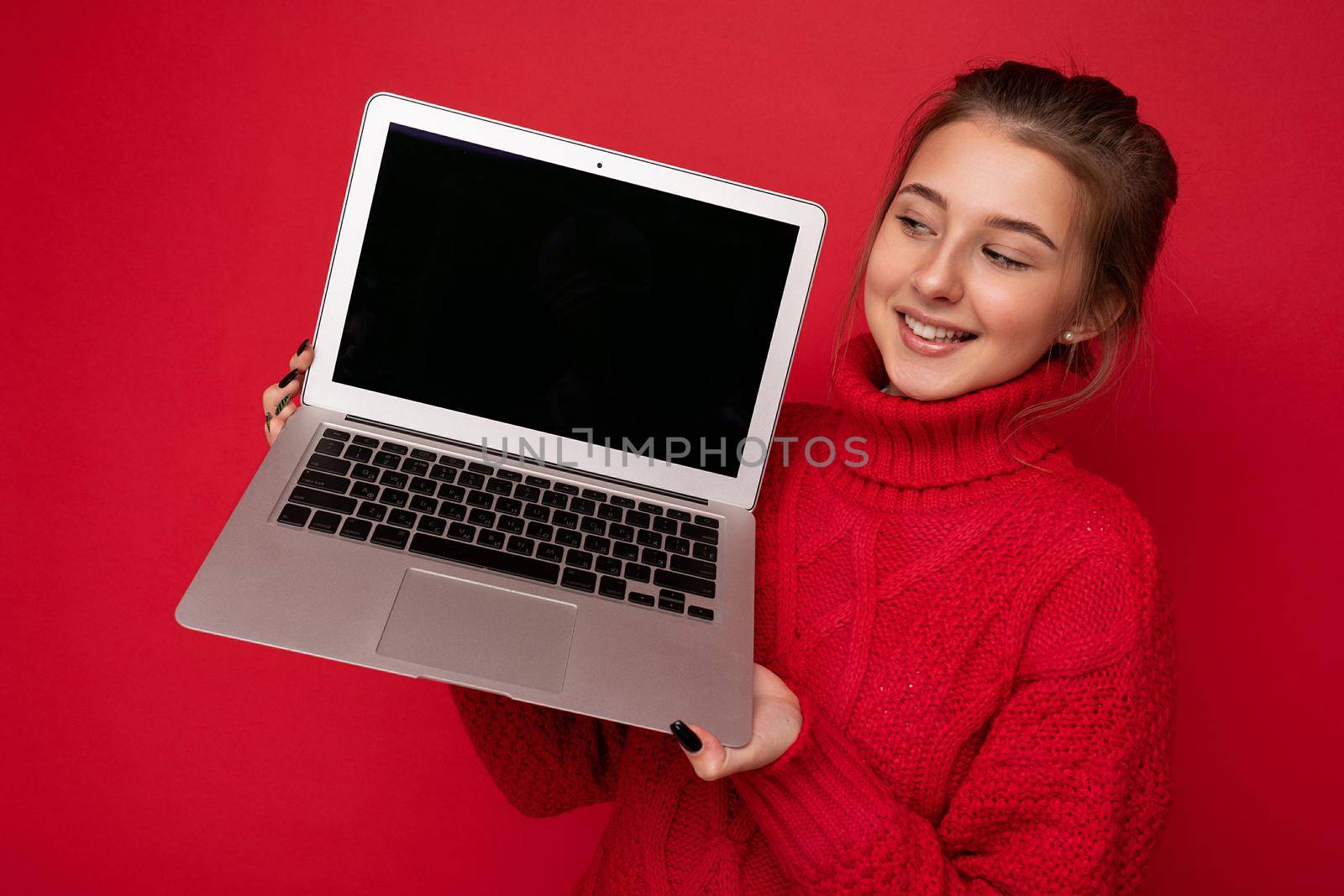 Photo of Beautiful satisfied happy young woman holding computer laptop looking at monitor display wearing red sweater isolated over red wall background. Copy space, mock up