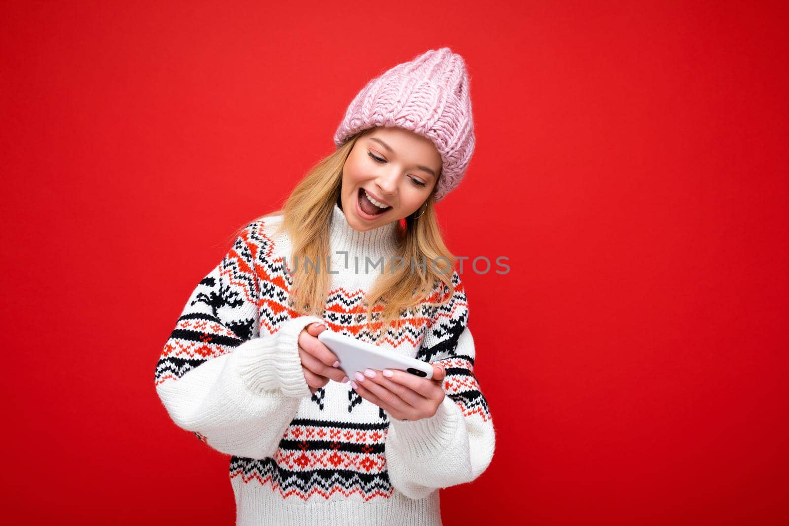 Photo of beautiful smiling young blonde woman wearing warm knitted hat and winter warm sweater standing isolated over red background playing games via smartphone looking at device display.