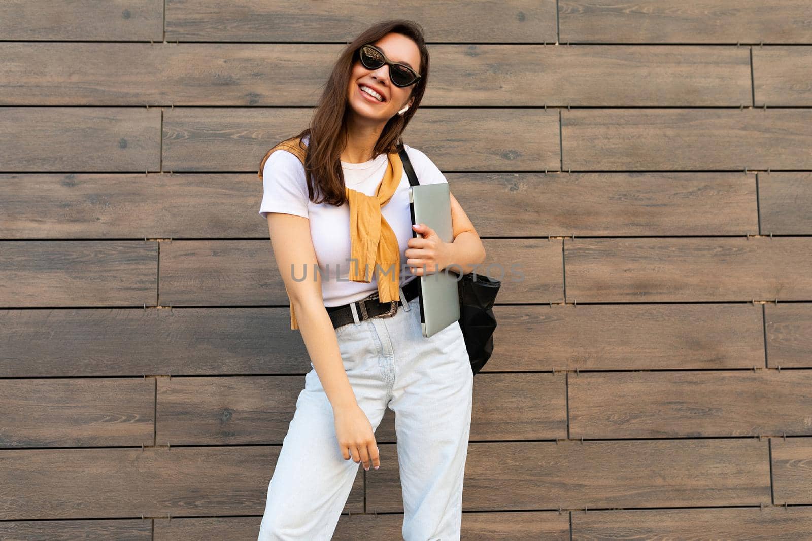 Fascinating beautiful smiling charming young brunet girl looking at camera holding computer laptop and black sunglasses in white t-shirt and light blue jeans in the street near brown wall.