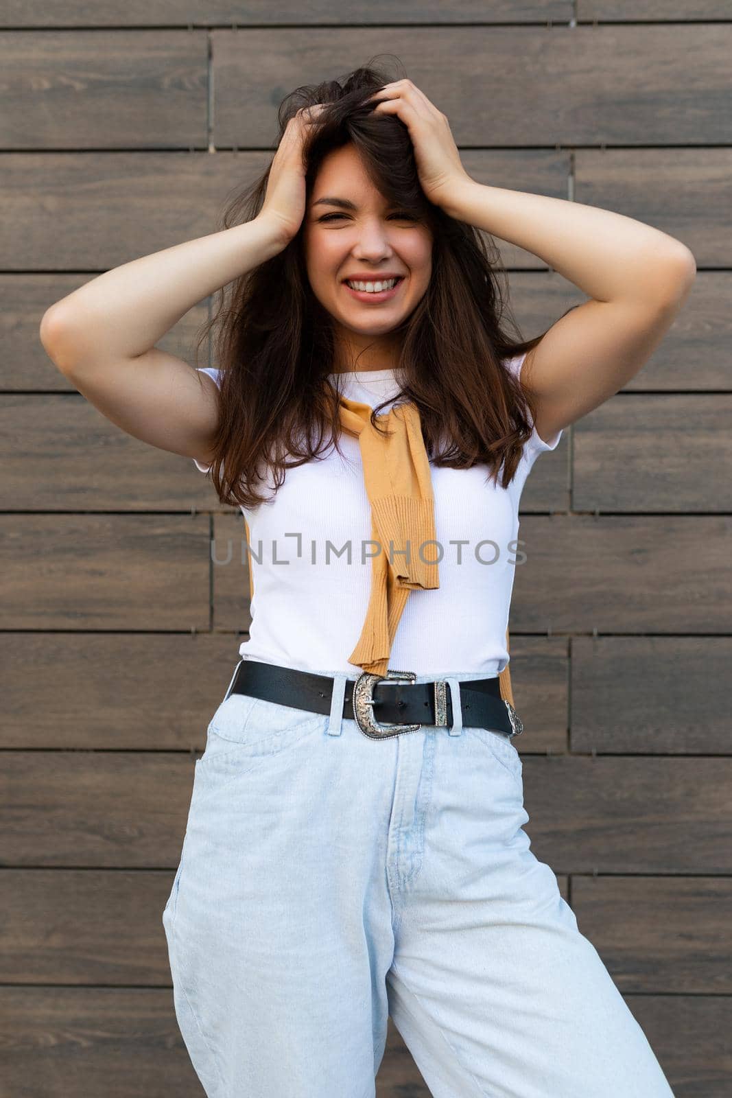 Vertical ptoto of young beautiful positive happy brunette woman standing against brown wall in the street and wearing stylish outfit.