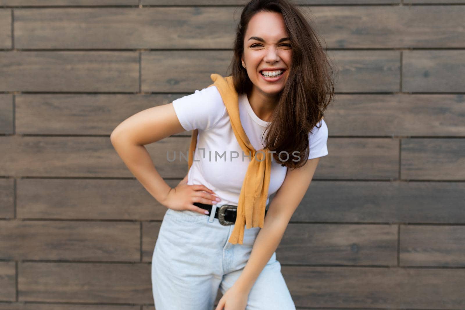 Portrait of successful smiling joyful happy young brunet woman wearing casual white t-shirt and jeans with yellow sweater poising near brown wall in the street and having fun.