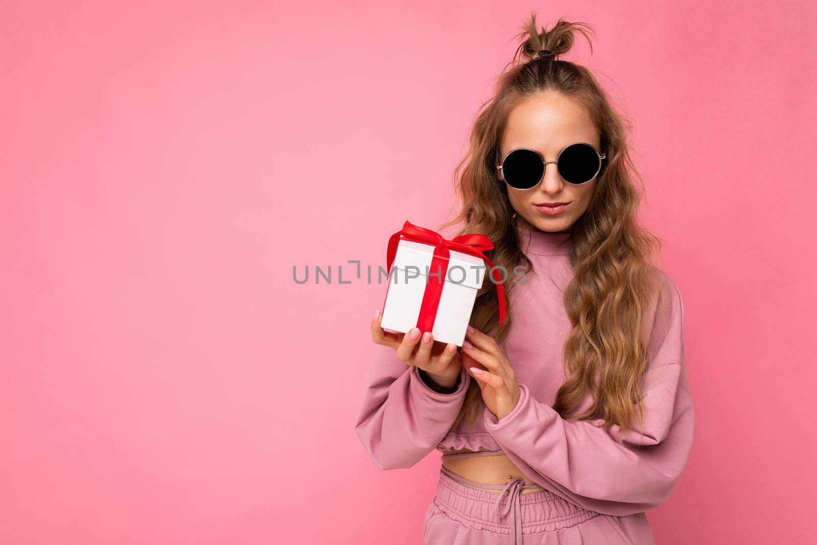 Photo of attractive happy self-confident young blonde curly female person isolated over pink background wall wearing pink sport clothes and sunglasses holding gift box looking at camera. Empty space