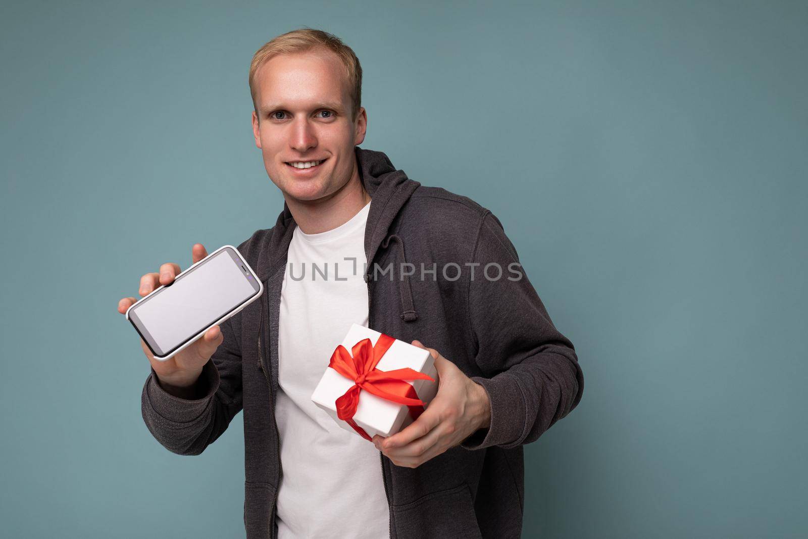 Photo shot of handsome happy smiling young man isolated over blue background wall wearing casual stylish clothes holding white gift box with red ribbon and mobile phone with empty screen display for mockup looking at camera.