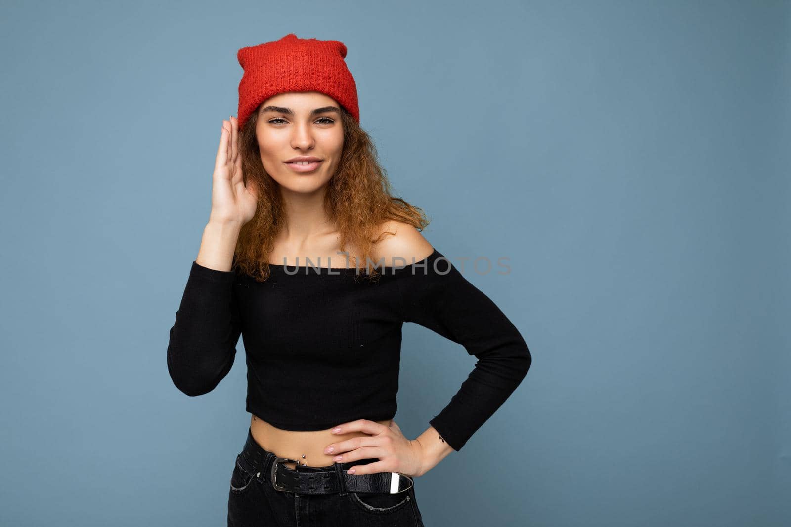 Photo of young positive happy beautiful brunette curly woman with sincere emotions wearing black crop top and red hat isolated on blue background with copy space and listening attentively.