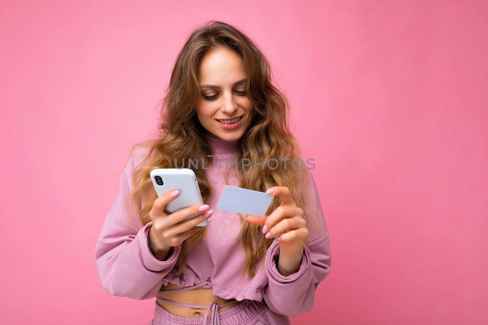 Attractive happy joyful young blonde curly woman wearing pink clothes isolated over pink background using mobile phone paying online shopping through credit card looking at smartphone screen.