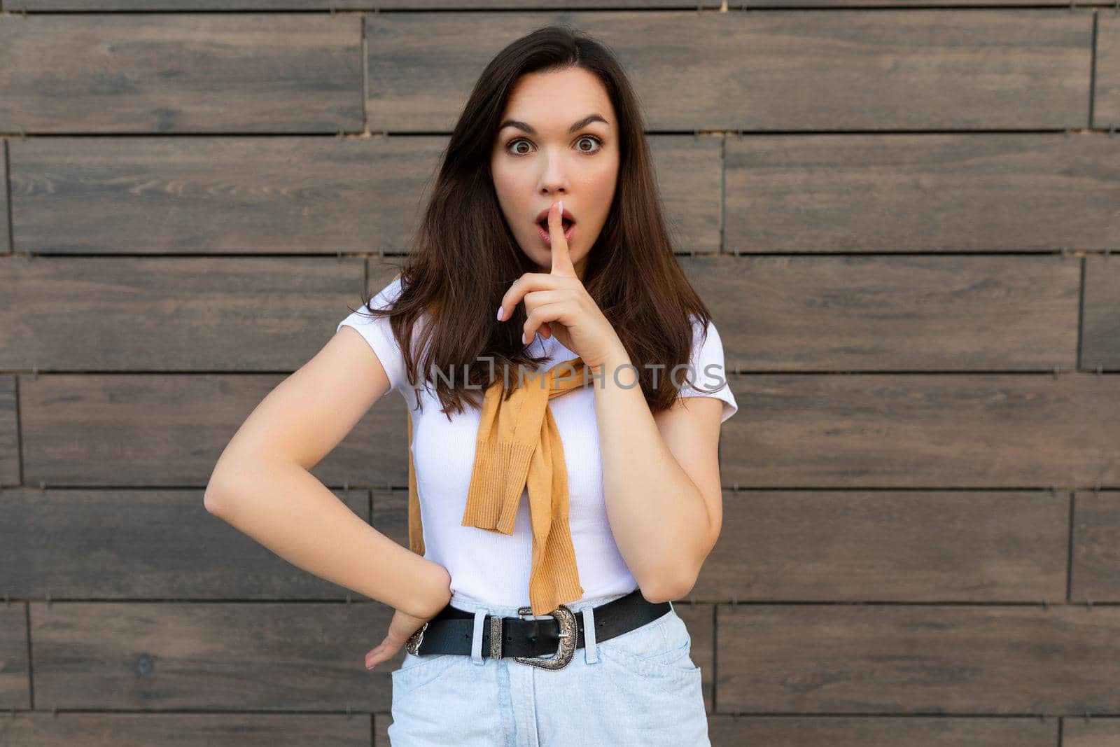 Photo of pretty young brunette woman in trendy outfit. Beautiful attractive female person standing outside in the street and showing shh gesture. Keep secret concept.