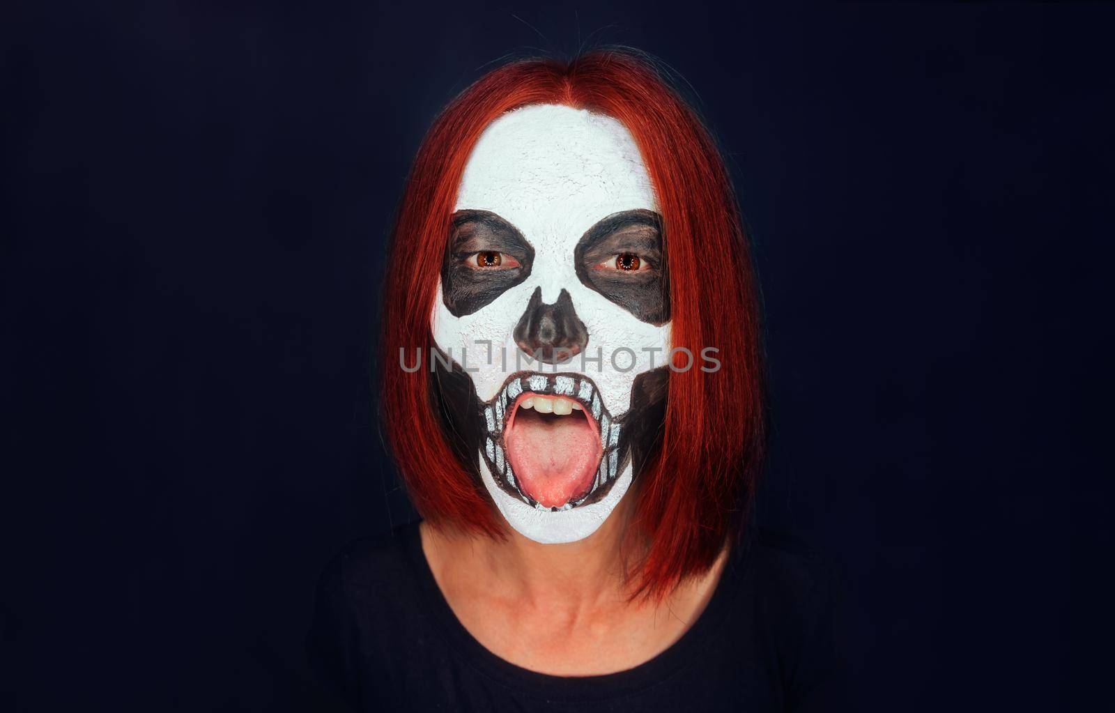 Woman with skull makeup and red hair showing tongue on dark background, Halloween or horror theme