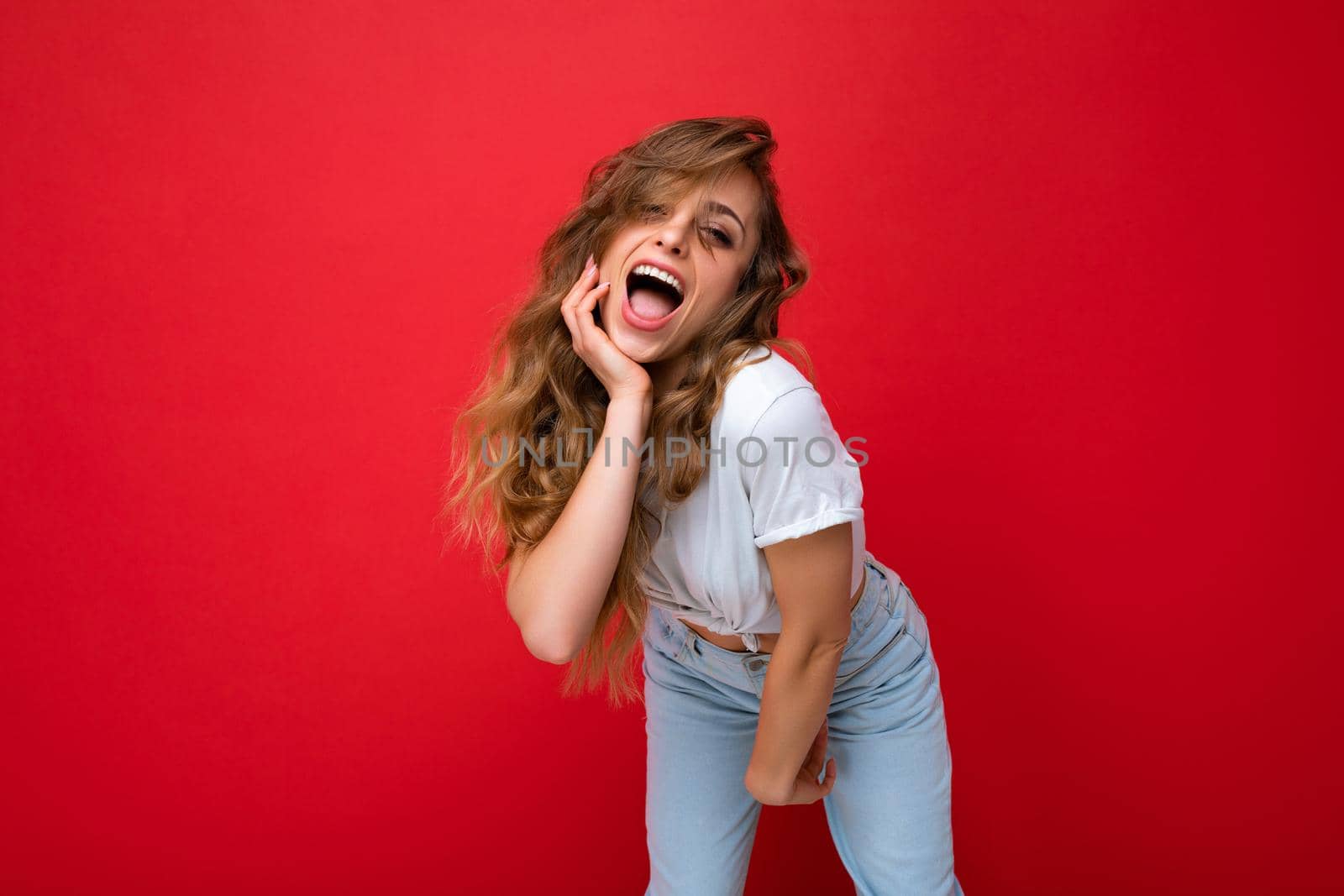 Young beautiful blond woman wearing white t-shirt looking at camera. Positive female shows facial sincere emotions. Funny model isolated on red wall with empty space.