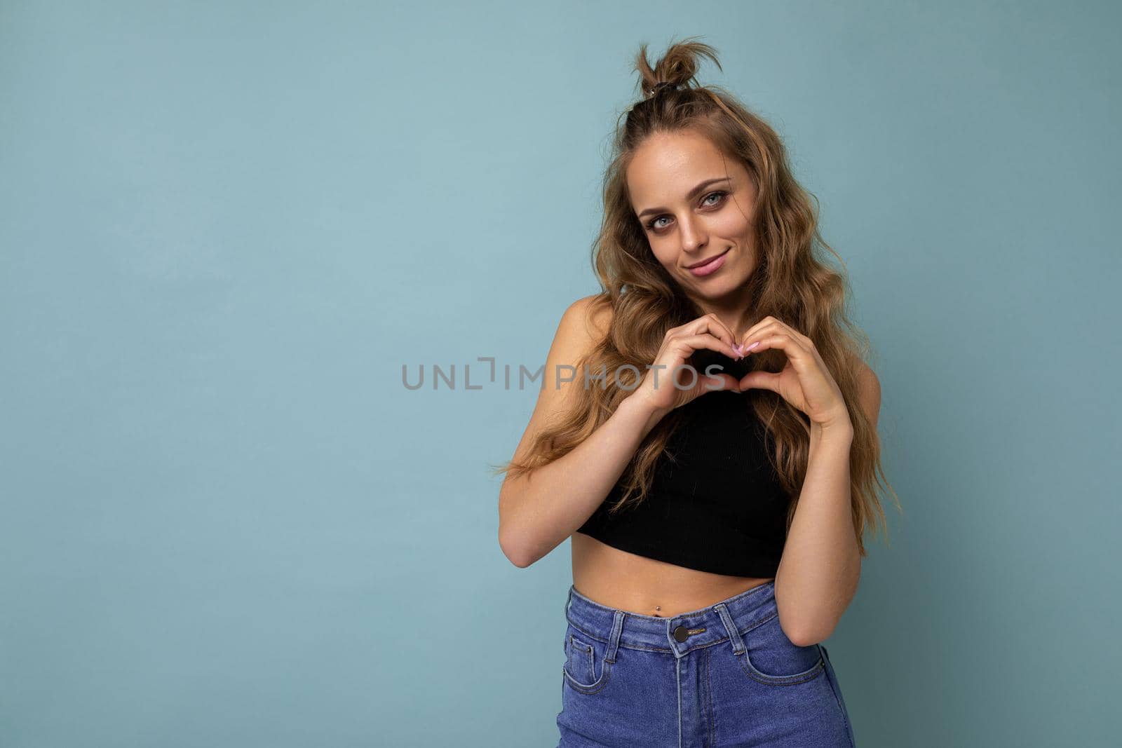Photo of young positive happy smiling beautiful woman with sincere emotions wearing stylish clothes isolated over background with copy space and showing heart gesture.