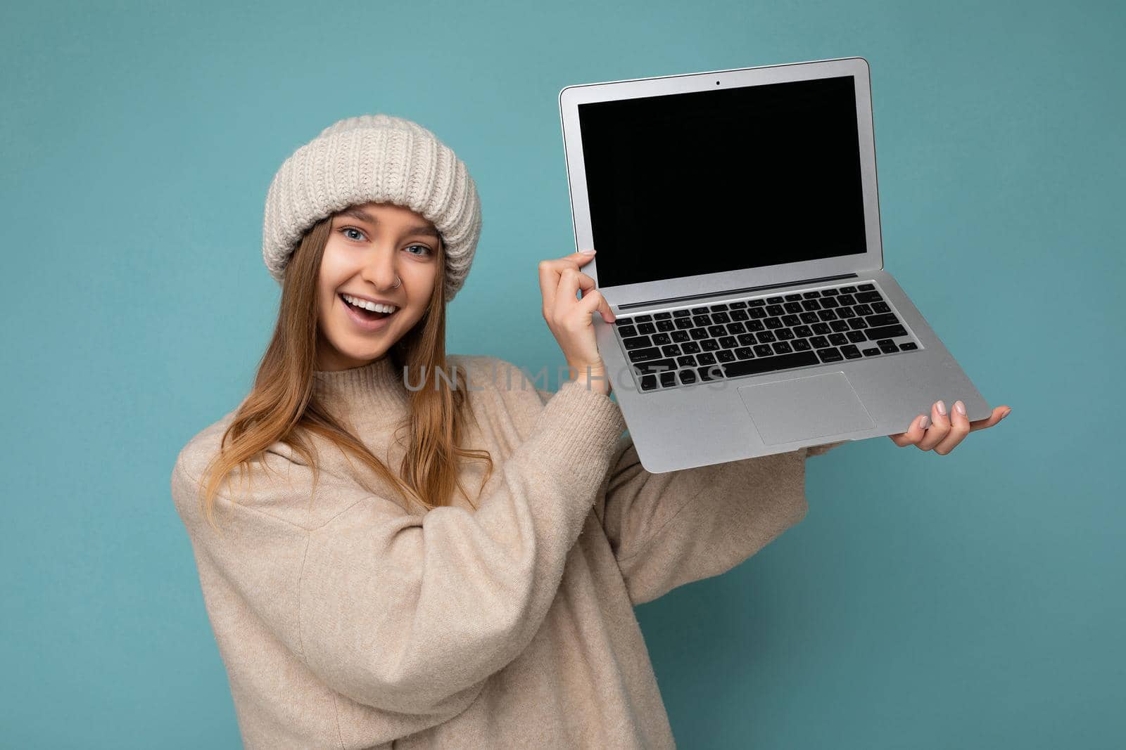 beautiful charming fascinating pretty smiling happy young dark blond feemale student woman in winter warm knitted beige hat holding computer laptop looking at camera wearing beige winter sweater isolated over light blue wall background in studio. Cutout, free empty space for advertising and advertisement