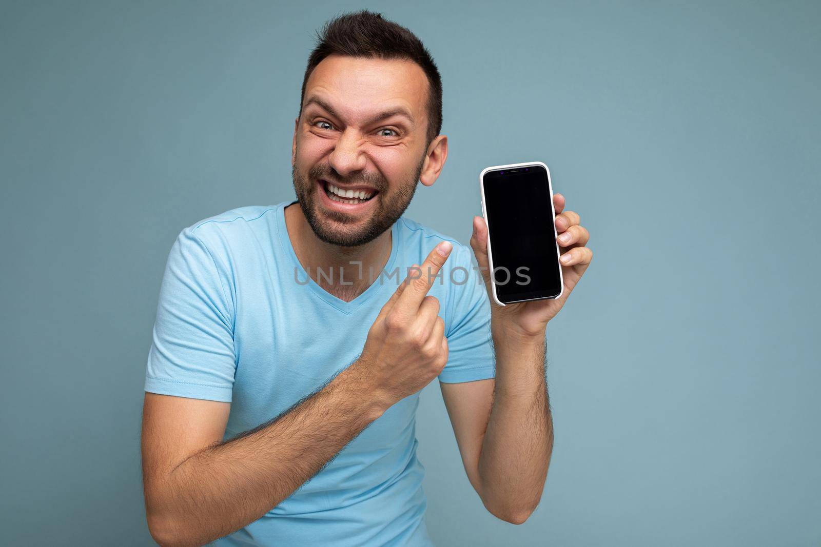 Photo of handsome crazy emotional smiling adult male person good looking wearing casual outfit standing isolated on background with copy space holding smartphone showing phone in hand with empty screen display for mockup pointing at gadjet looking at camera.