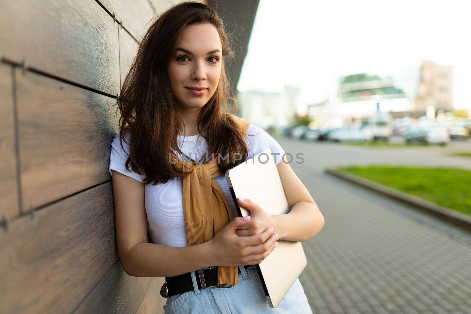 beautiful nice-looking charming attractive lovely fascinating cheerful cheery straight-haired brunet woman looking at camera in the street with laptop computer in hands with white t-shirt and blue jeans.