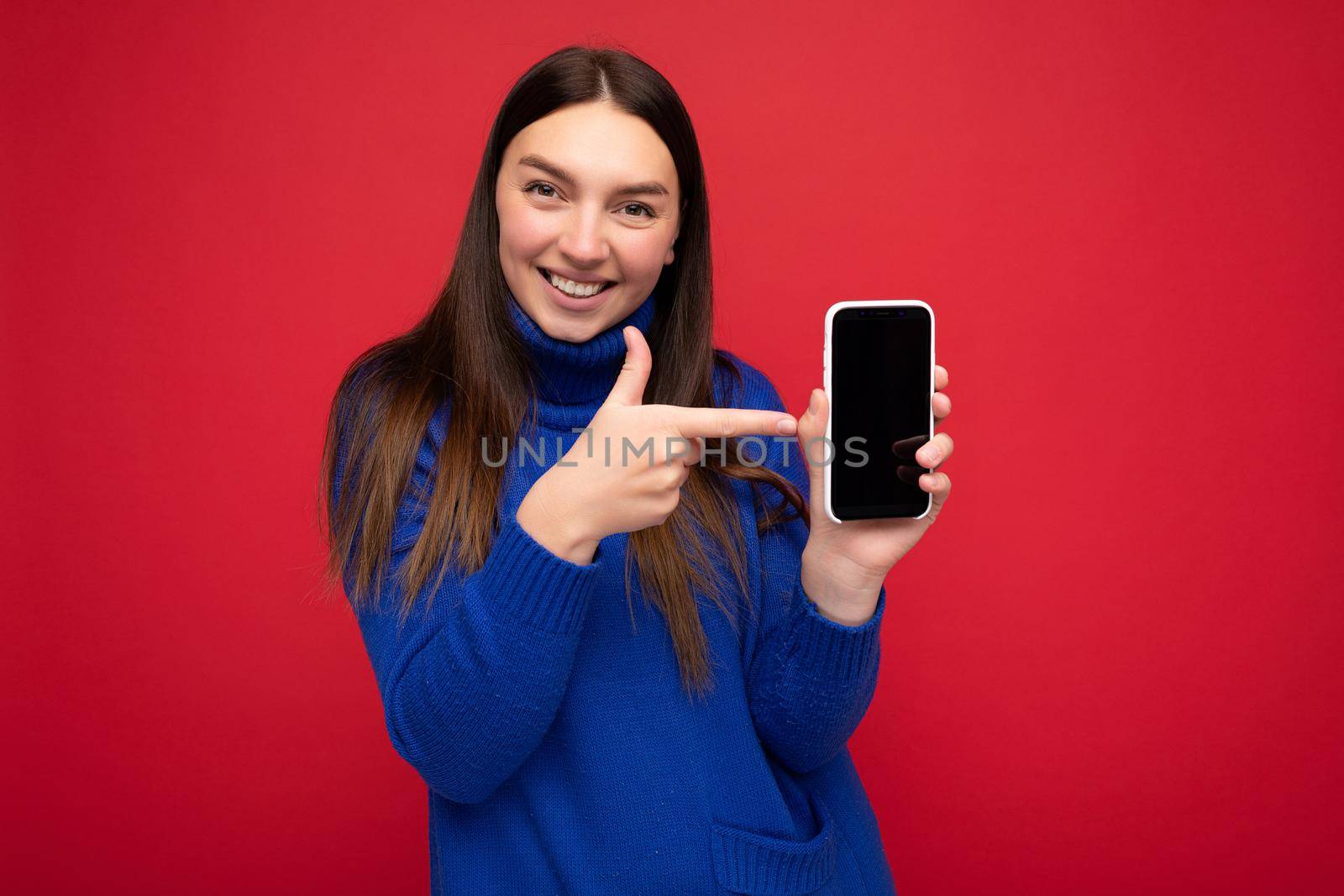 Photo of beautiful smiling young woman good looking wearing casual stylish outfit standing isolated on background with copy space holding smartphone showing phone in hand with empty screen display for mockup pointing at gadjet looking at camera.