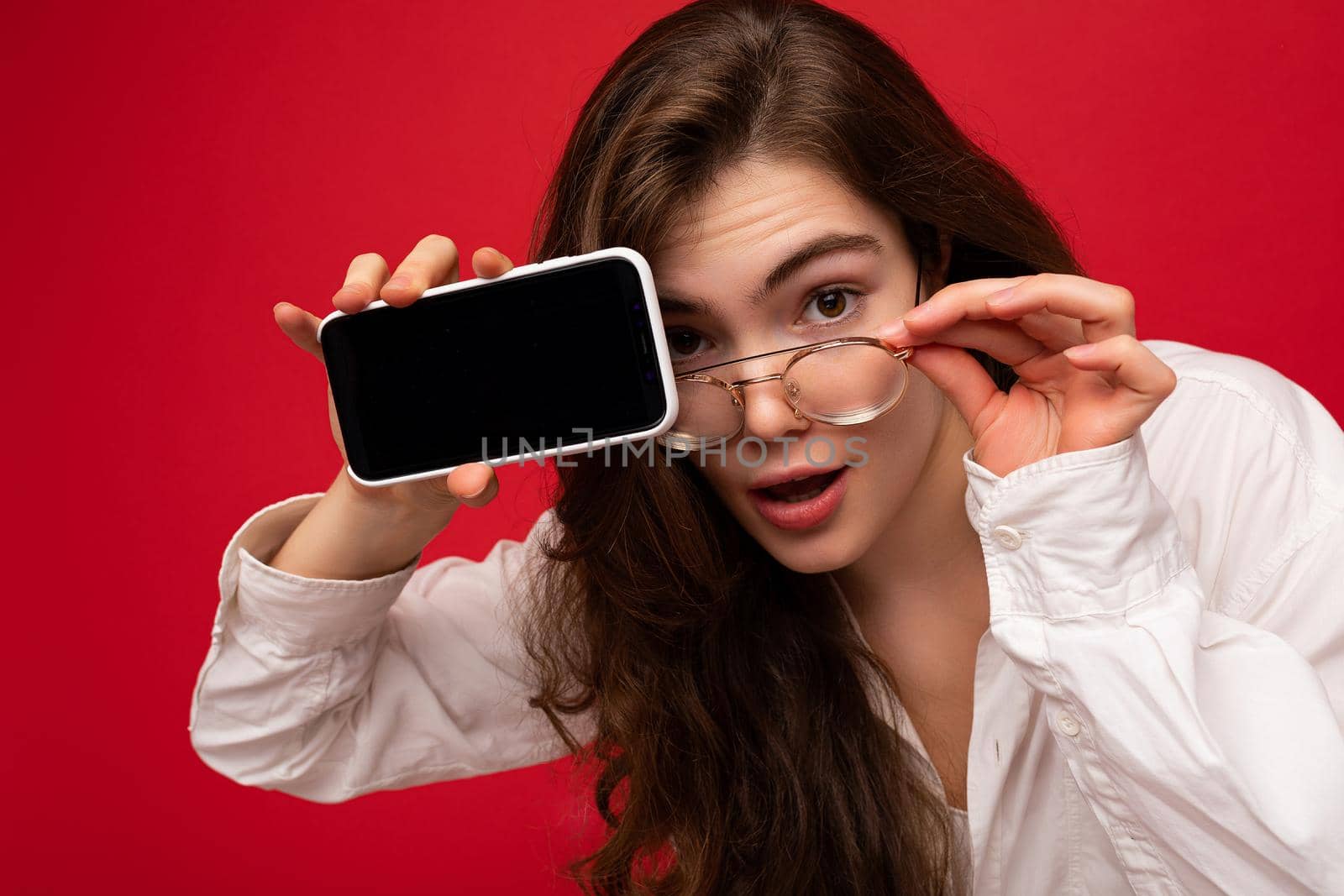 beautiful young brunette woman wearing white shirt and optical glasses isolated over red background holding in hand and showing mobile phone with empty screen for cutout looking at camera.