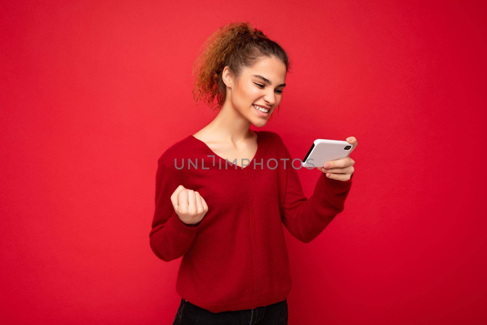 Young emotional happy woman wearing dark red sweater isolated over red background holding smartphone having fun and showing yes gesture looking at mobile phone screen.