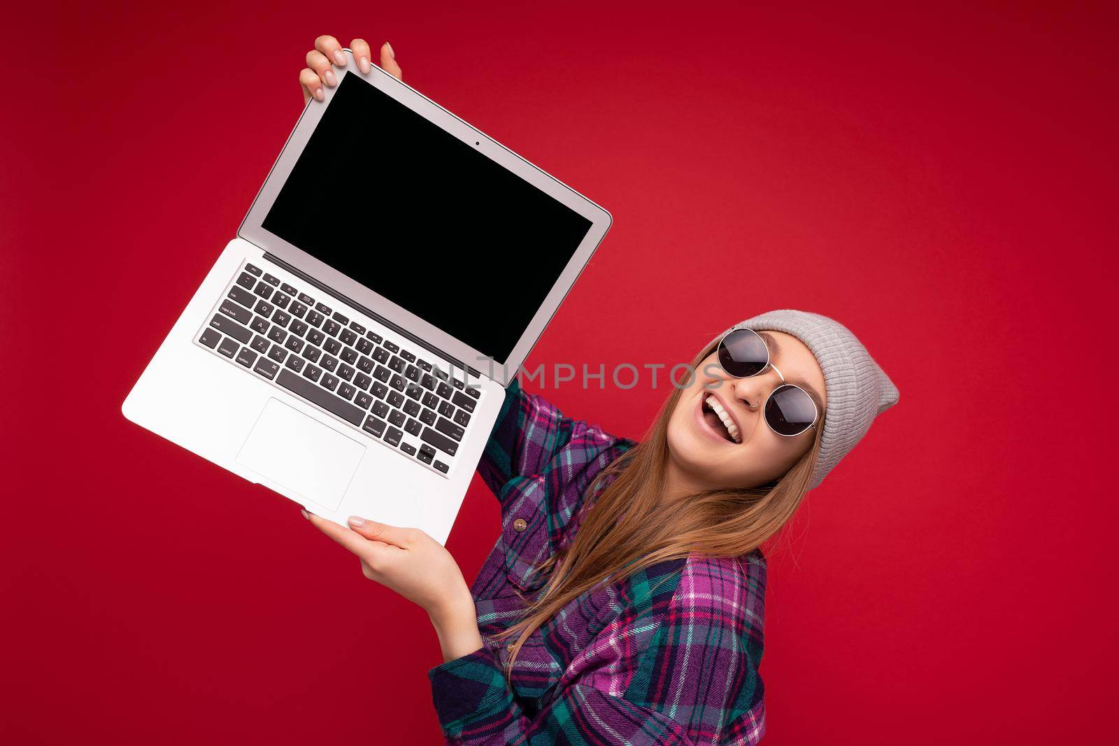 Photo shot of beautiful smiling dark blond young feemale teenager holding computer laptop with empty monitor screen with mock up and copy space wearing sun glasses hat and colourful shirt looking at camera isolated over red wall background.