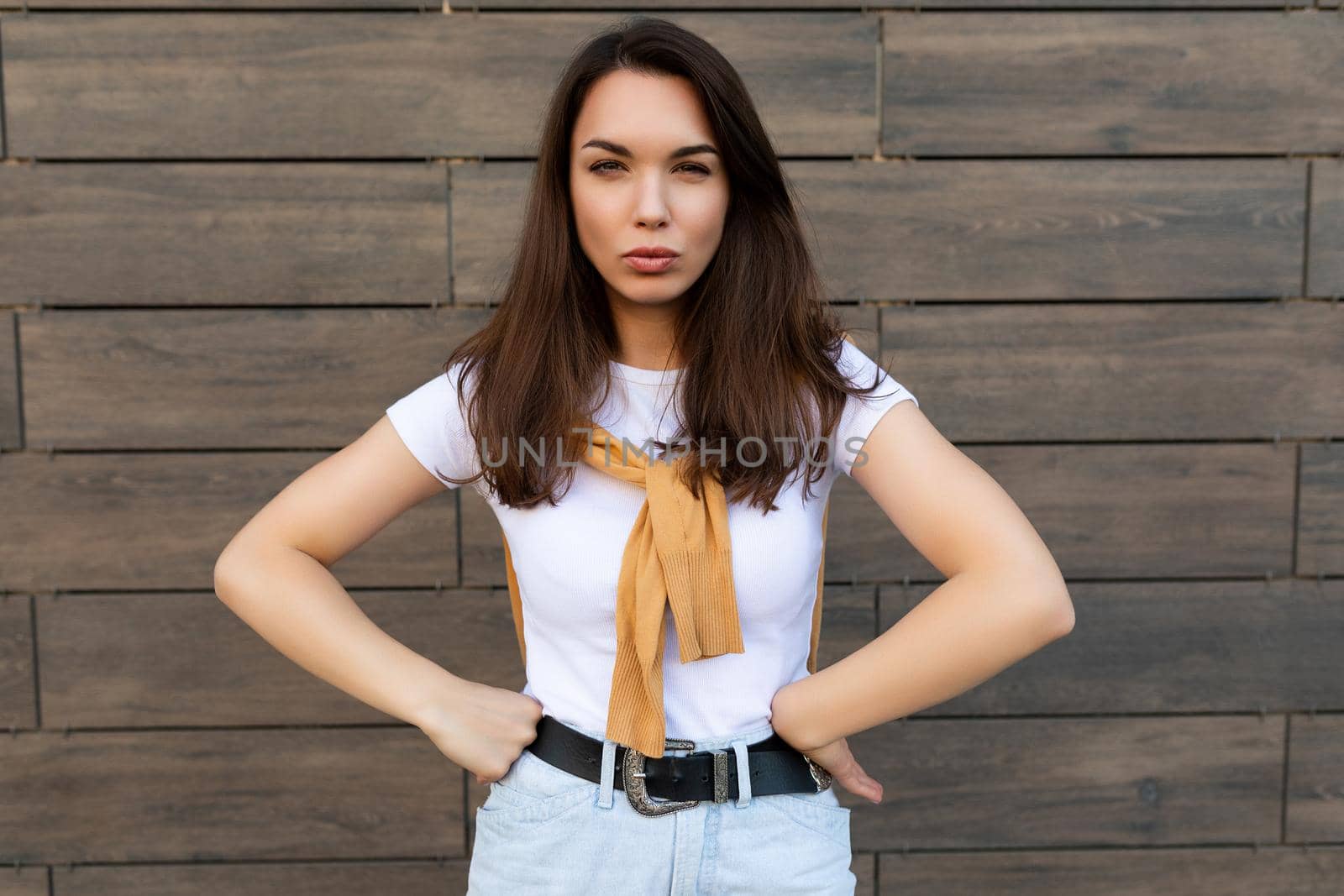 Portrait of touchy sad upset offended young brunet woman wearing casual white t-shirt and jeans with yellow sweater poising near brown wall in the street.