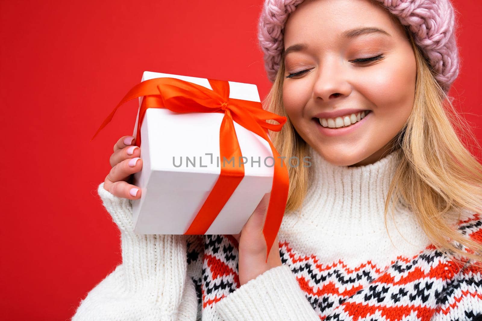 Portrait photo shot of beautiful happy smiling young blonde woman isolated over red background wall wearing winter sweater and pink hat holding white gift box with red ribbon and having fun.