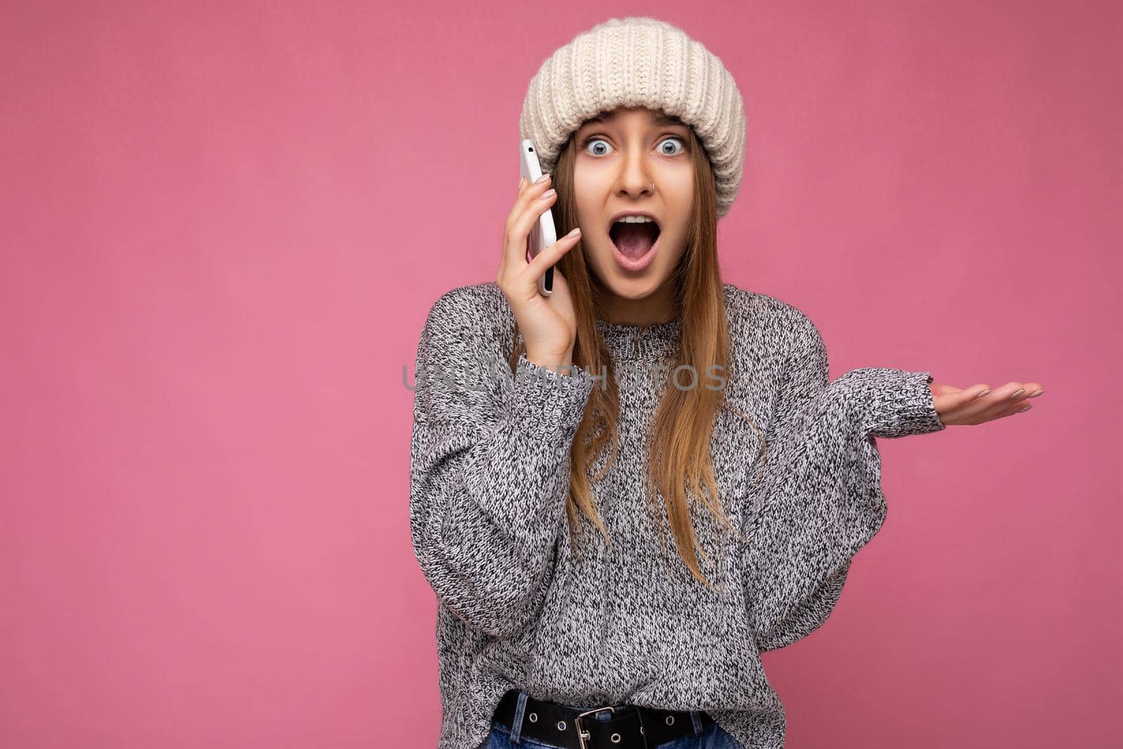 Closeup portrait of Attractive amazed young blonde woman wearing casual grey sweater and beige hat isolated over pink background holding in hand and talking on mobile phone looking at camera with open mouth.