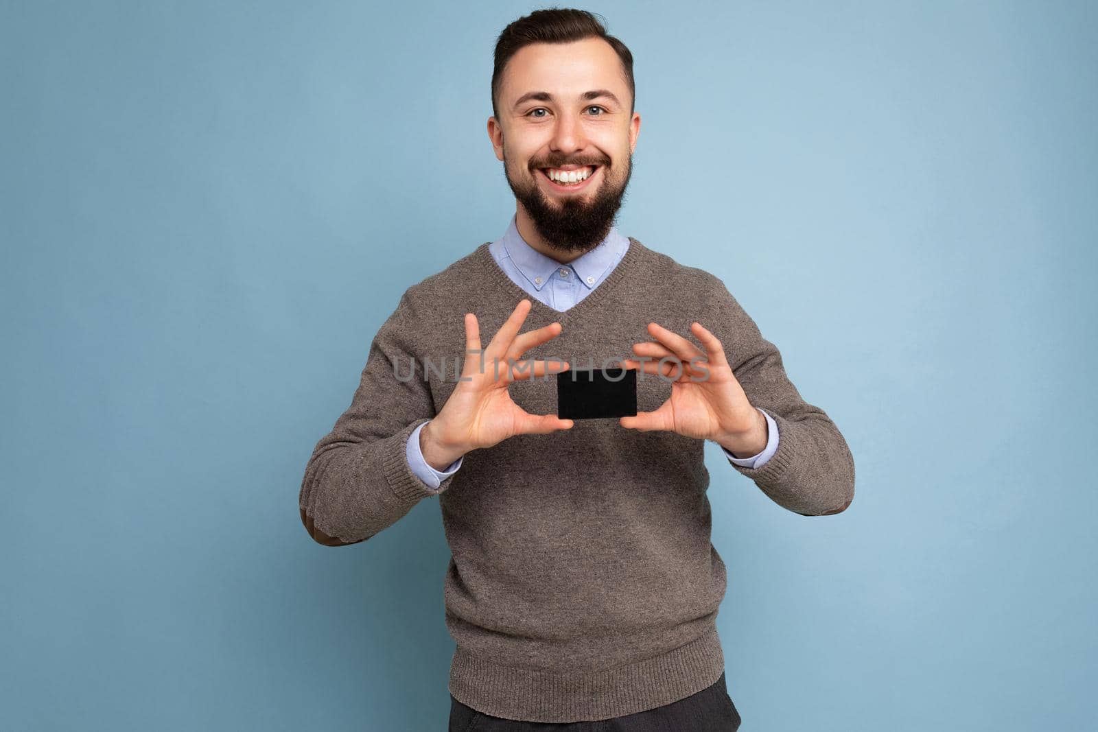 Handsome joyful smiling brunette bearded young man wearing grey sweater and blue shirt isolated on background wall holding credit card looking at camera.