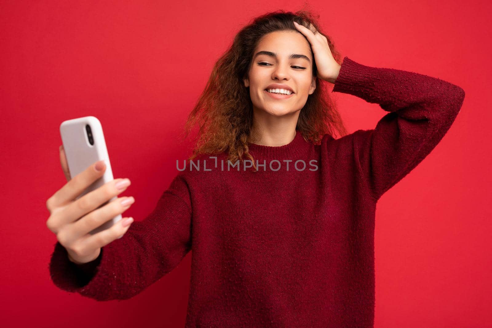 Smiling happy Beautiful young woman with curly hair wearing dark red sweater isolated on red background wall holding and using smart phone looking at telephone screen and taking selfie.