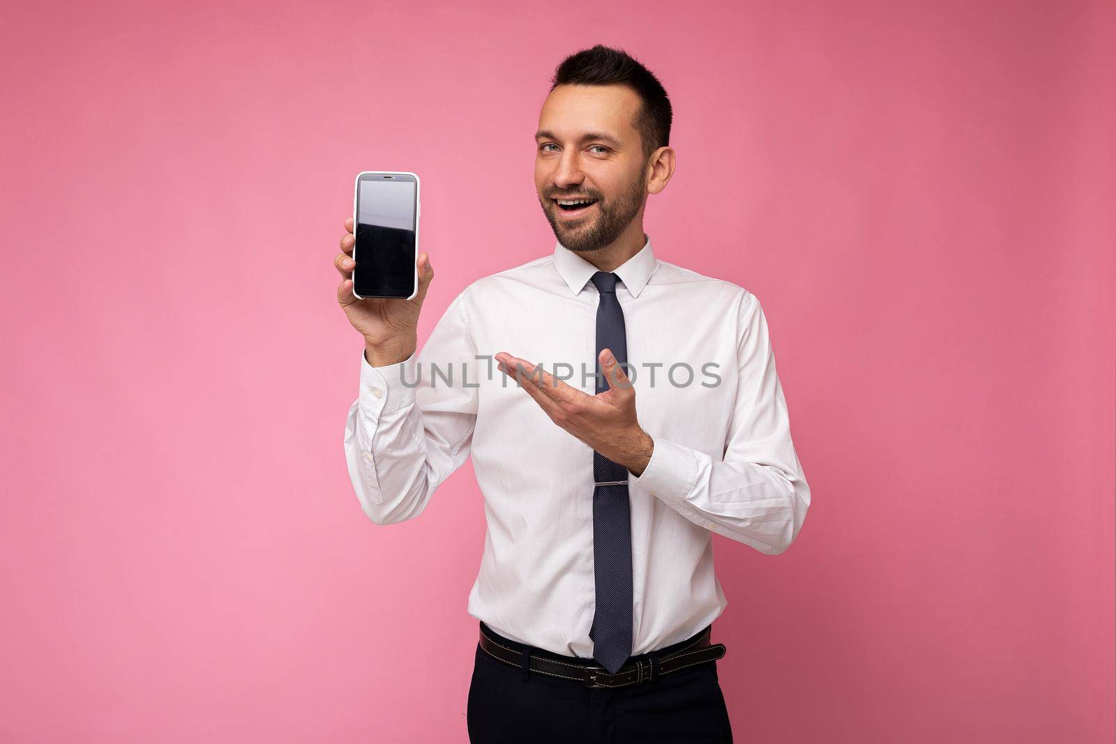 Photo of handsome smiling adult male person good looking wearing casual outfit standing isolated on background with copy space holding smartphone showing phone in hand with empty screen display for mockup pointing at gadjet looking at camera.